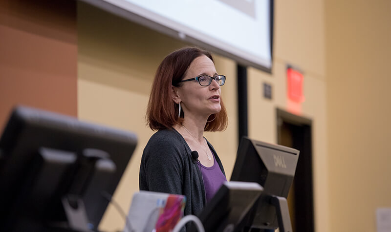 A woman in glasses stands at a podium in front of a screen at North Dakota University Systems.