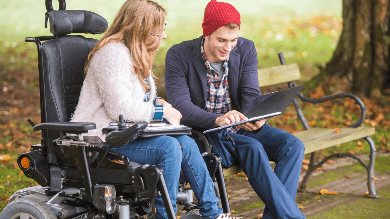 A man in a red hat looking at a laptop with a woman in a wheelchair.